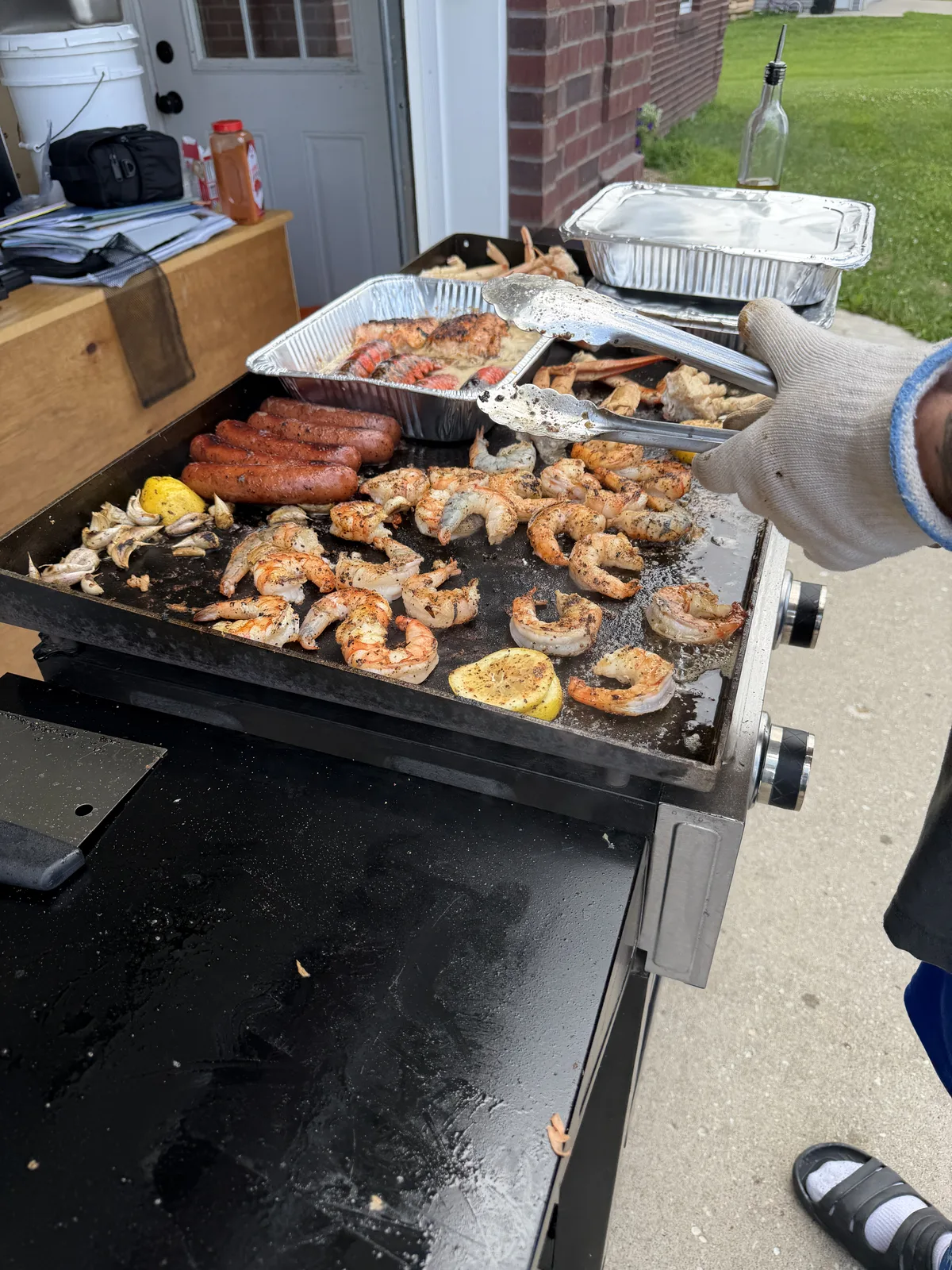 Overhead view of the Blackstone griddle loaded with shrimp, sausage, crab, and a foil pan, with tongs plating into a tray