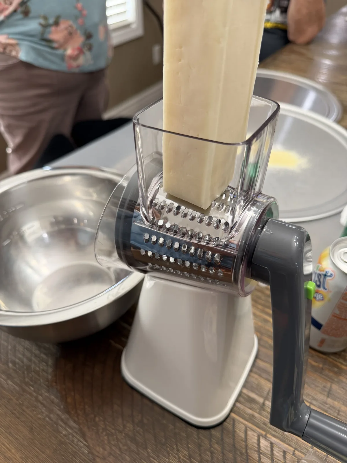 A block of mozzarella being fed through a hand-crank rotary grater into a metal bowl