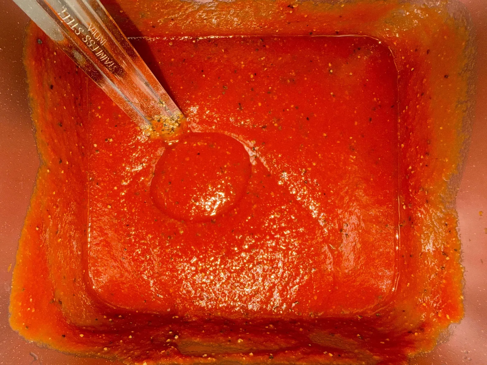 Seasoned pizza sauce stirred in a stainless steel container, flecked with dried herbs