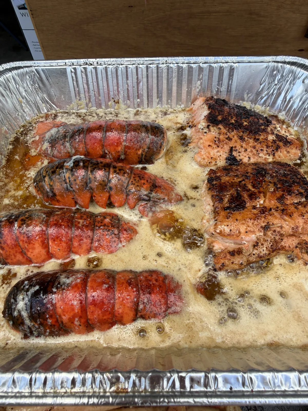 Four lobster tails and two seared salmon fillets bubbling in a foil pan of butter