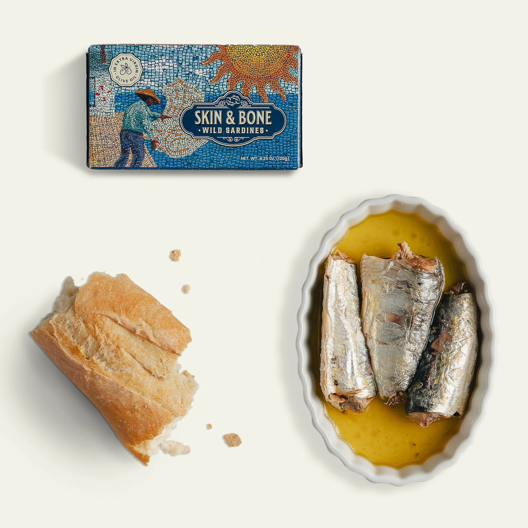 A box of Skin and Bone wild caught sardines beside a torn piece of crusty bread and an oval white dish holding three sardine fillets in golden olive oil