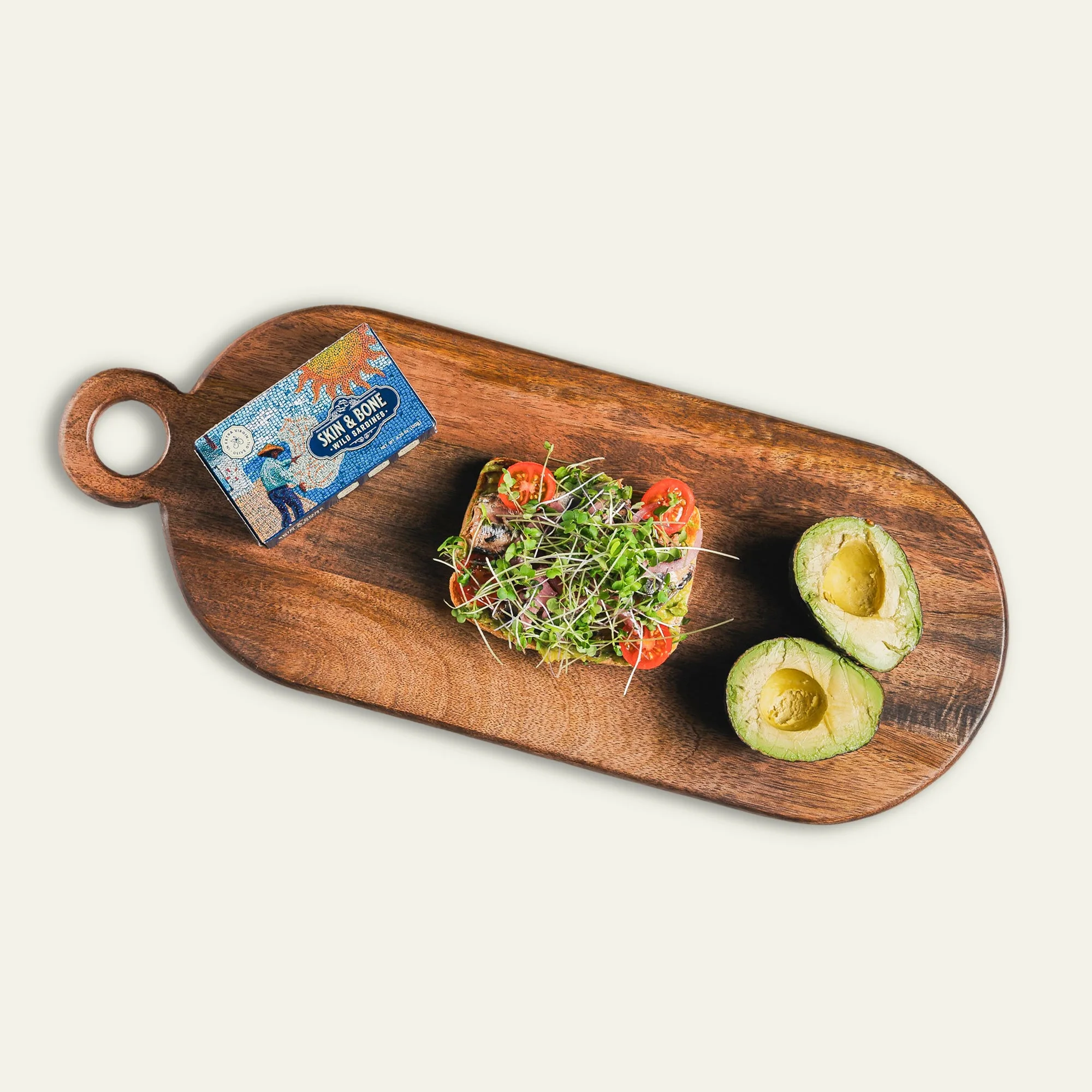 A wooden serving board with a tin of Skin and Bone sardines, an avocado halved with the pit, and toast topped with sardines, cherry tomatoes, and microgreens