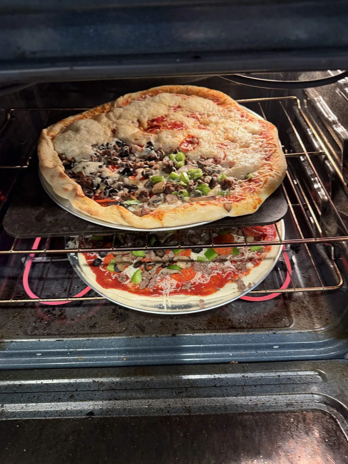 Two homemade pizzas on pans in the oven, one already golden and one still raw and freshly topped